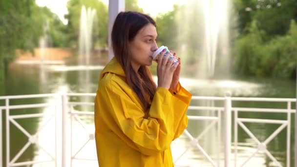 Una chica con un impermeable amarillo bebe café. Fuentes en un parque verde. — Vídeo de stock