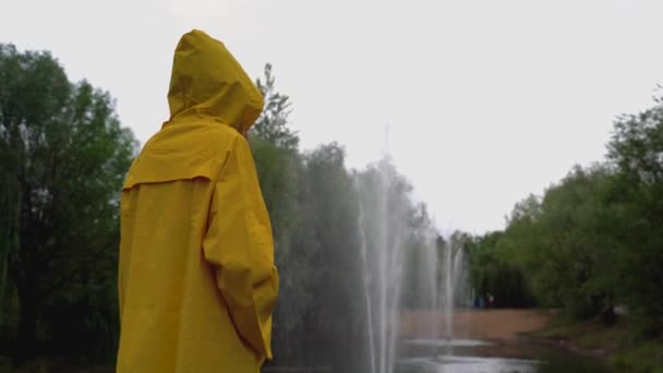 Дівчина в жовтому плащі дивиться на ставок з фонтанами в парку . — стокове відео