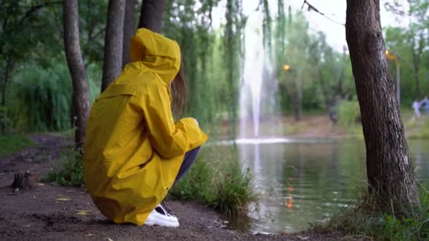 Ein Mädchen im gelben Regenmantel sitzt im Park und wirft Steine ins Wasser. — Stockvideo