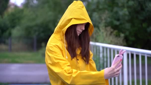 En flicka i gul regnrock gör en selfie på en smartphone i parken. — Stockvideo