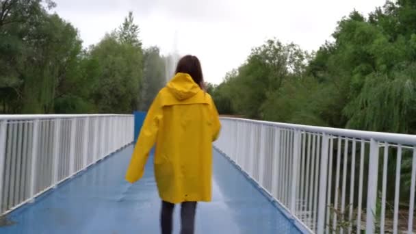 Uma menina de capa amarela bebe café e caminha ao longo da ponte. — Vídeo de Stock