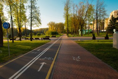 Güneşli bahar sabahının erken saatlerinde boş bisiklet yolu altyapısı. Parkta kimse yok, yeşil çimenler, ağaçlar ve mavi gökyüzü. Mutlu sıcak ışık ve parlak renkler.