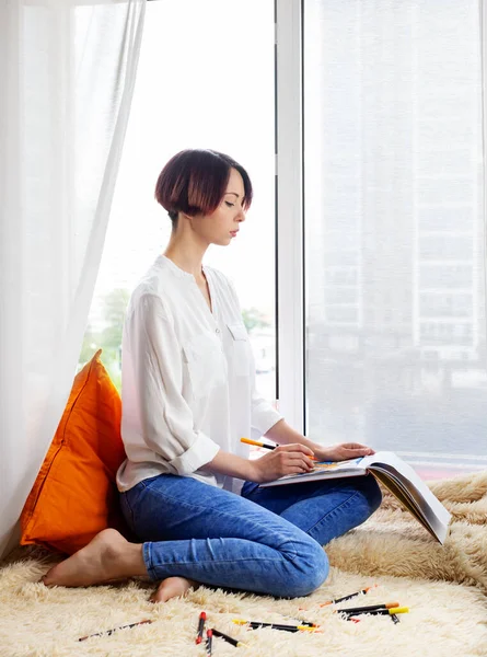 Sad young woman drawing with sketch markers in her sketcbook near the window on yellow furry plaid with orange pillows. Interesting hobby at home, leisure during social distancing. Vertical, copy space.