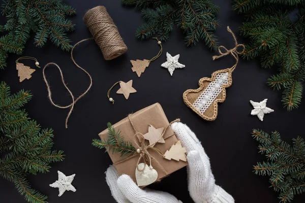 Sıfır atık Noel dairesi elleri beyaz eldivenlerle uzanmış siyah arka planda kraft kağıdında bir hediye tutuyordu. Yeniden kullanılabilir kağıt, sicim, el yapımı dekor. Çevre dostu ev yapımı yeni yıl hediyesi.