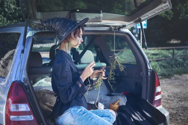 Halloween party and social distancing. Lonely young woman in a witch costume and bright makeup holding dry herbs in trunk of car with halloween decor. Trick-or-treats. Alternative safe celebration