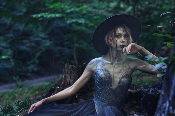 Halloween Witch in a dark magic forest. Beautiful young woman dressed in witches hat and creative costume with bright makeup. Halloween social distancing. Alternative safe celebration