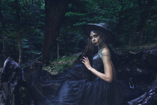 Halloween Witch in a dark magic forest. Beautiful young woman dressed in witches hat and creative costume with bright makeup. Halloween social distancing. Alternative safe celebration