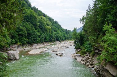 Dağlardaki nehir. Karpatya kırsalının harika bahar manzarası. Orman ve kayalık kıyılar arasında mavi yeşil su. Gökyüzünde bulutlarla güneşli bir gün.