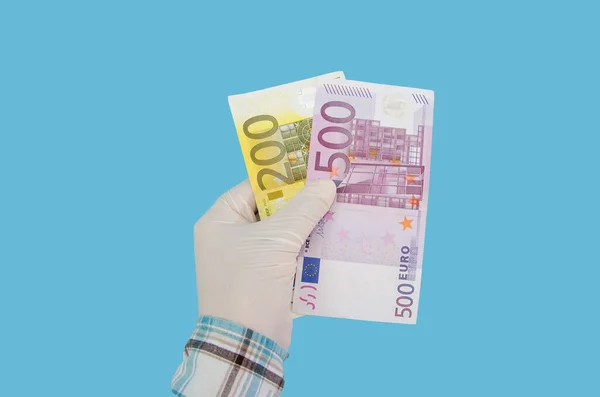 hand in a white medical glove with euro banknotes isolated on a blue background. 200 and 500 euros in a woman\'s hand.