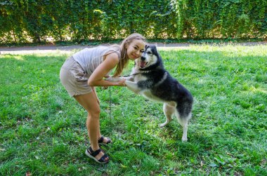 Çekici bir sarışın köpeğini pençelerinden tutup kameraya bakar. Parkta köpeği olan kız.