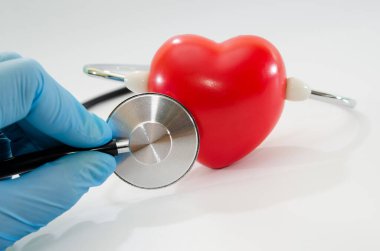 Beyaz arka planda steteskopla kırmızı kalbi inceleyen lateks eldivenli doktor görüntüsü.