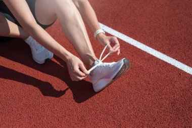 Spor ayakkabı bağcıklarını eğitirken bağlayan kadın. Spor kıyafetli bir kadın ayakkabı bağı bağlayıp çömeliyor. Açık hava eğitim stadyumunda gri gardırop ve beyaz spor ayakkabılar, kadın vücudunun yakın çekimi.