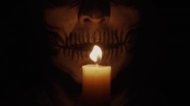 İskelet makyajlı bir adam kameraya bakar. Adam kızgın ve bir büyü yapıyor. Mumların loş ışığında iskelet. Cadılar Bayramı ve korku konsepti.