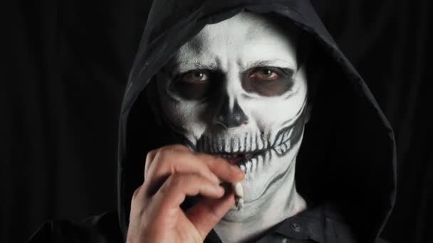 有化妆骷髅和黑色头罩的男人在黑暗的背景 骷髅抽一支香烟 吸烟使人死亡 万圣节或恐怖主题 优质4K视频 — 图库视频影像
