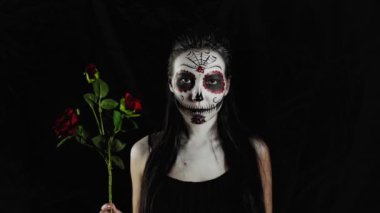 Cadılar Bayramı maskesi Calavera Catrina. Elinde gül olan bir kız. Meksika Ölüler Günü. Karanlık bir arka planda cadılar bayramı için korkunç renkli makyajlı genç bir kadının portresi. 4K. 