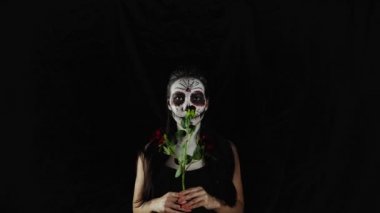 Cadılar Bayramı maskesi Calavera Catrina. Elinde gül olan bir kız. Meksika Ölüler Günü. Karanlık bir arka planda cadılar bayramı için korkunç renkli makyajlı genç bir kadının portresi. 4K. 