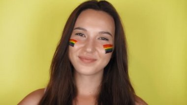 Yüzünde gökkuşağı deseni olan mutlu bir kadın. Yüzünde LGBT bayrağı var. Sarı arkaplan Yüksek kalite 4k video