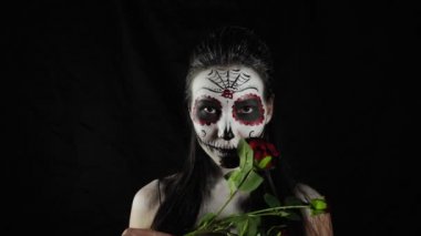 Cadılar Bayramı maskesi Calavera Catrina. Elinde gül olan bir kız. Meksika Ölüler Günü. Karanlık bir arka planda cadılar bayramı için korkunç renkli makyajlı genç bir kadının portresi. 4K. 