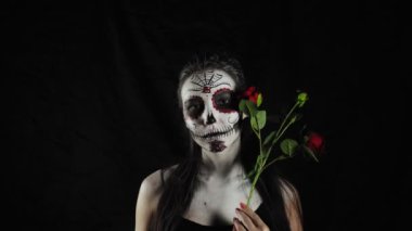 Cadılar Bayramı maskesi Calavera Catrina. Elinde gül olan bir kız. Meksika Ölüler Günü. Karanlık bir arka planda cadılar bayramı için korkunç renkli makyajlı genç bir kadının portresi. 4K. 