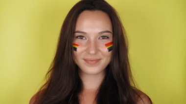 Yüzünde gökkuşağı deseni olan mutlu bir kadın. Yüzünde LGBT bayrağı var. Sarı arkaplan Yüksek kalite 4k video