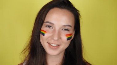 Yüzünde gökkuşağı deseni olan mutlu bir kadın. Yüzünde LGBT bayrağı var. Sarı arkaplan Yüksek kalite 4k video