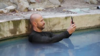 Bir adam dağlarda kaplıcanın tadını çıkarır. Genç bir adam havuzda bir telefon kullanır. Sağlık için sülfürik kaplıcalar.