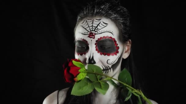 Halloween-Maske Calavera Catrina. Mädchen mit einer Rose in der Hand. Mexikanischer Tag der Toten. Porträt einer jungen Frau mit schrecklich buntem Make-up für Halloween auf dunklem Hintergrund. 4K. 