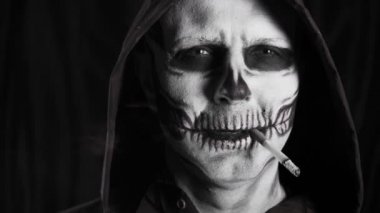 Siyah kapüşonlu ve esmer makyajlı bir adam. İskelet sigara içiyor. Sigara öldürür. Cadılar Bayramı ya da korku teması. Yüksek kalite 4k video