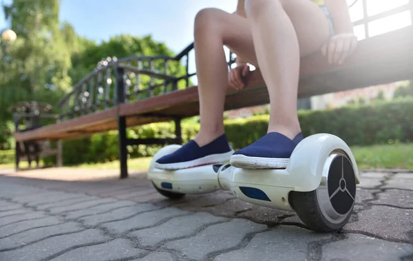 Los pies de una niña en un hoverboard mientras se relaja en un banco en un parque —  Fotos de Stock