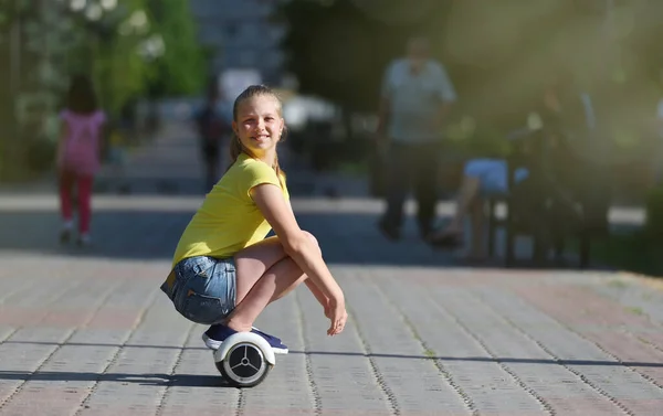 Meisje kind rijdt terwijl zitten op een hoverboard in een zomer park, gelukkig kijkend naar de camera — Stockfoto
