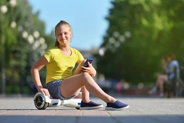 Usmívající se dívka dítě drží smartphone a sedí na hoverboard v pozadí zeleného parku a dívá se do kamery — Stock fotografie