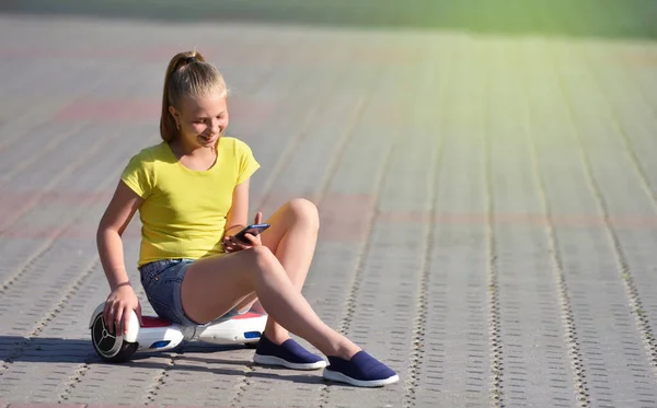 Ευτυχισμένο παιδί κορίτσι διαβάζει αυθεντικά μηνύματα σε ένα smartphone, ενώ περπατά σε ένα πάρκο σε ένα hoverboard, αυθεντική φωτογραφία — Φωτογραφία Αρχείου
