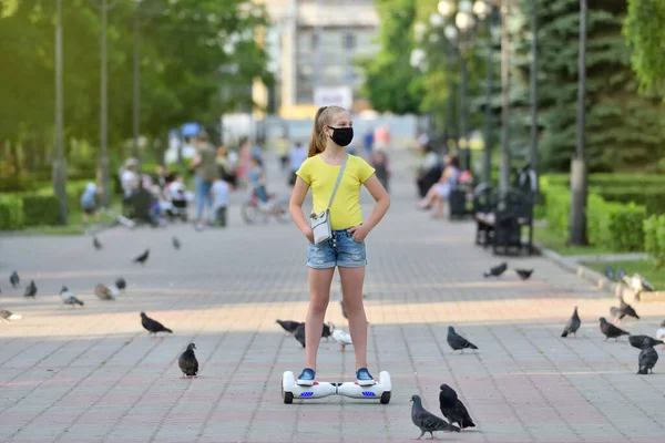 Dívka dítě v černé ochranné masce jezdí na vznášedle kolem holubů v parku během pandemie COVID-19 — Stock fotografie