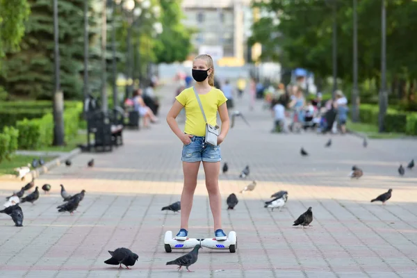 Anak perempuan bertopeng hitam pelindung menunggangi sebuah hoverboard melewati merpati di taman selama pandemi COVID-19 — Stok Foto