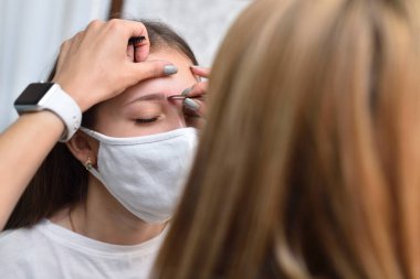 18-20 yaşlarındaki genç bir kız güzellik salonunda koruyucu bir maske takıyor ve salgın sırasında kaşlarını düzeltiyor.