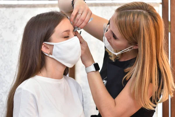 Ein junges Mädchen mit Schutzmaske korrigiert ihre Augenbrauen in einem Schönheitssalon, soziale Distanzierung während der COVID-19-Pandemie — Stockfoto