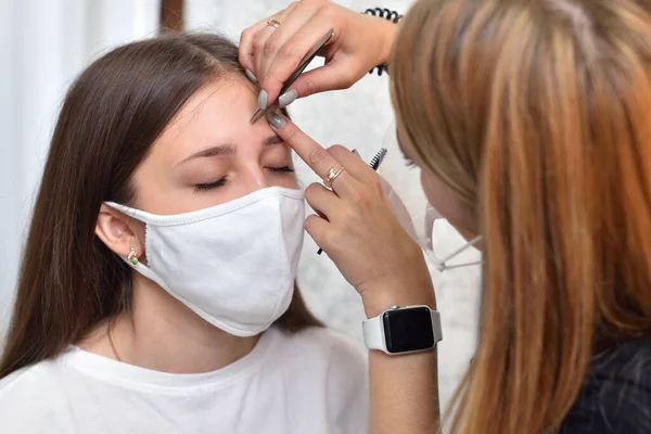 Uma jovem com uma máscara protetora branca em um salão de beleza arranca as sobrancelhas antes de colorir, distanciamento social durante a pandemia de COVID-19 — Fotografia de Stock