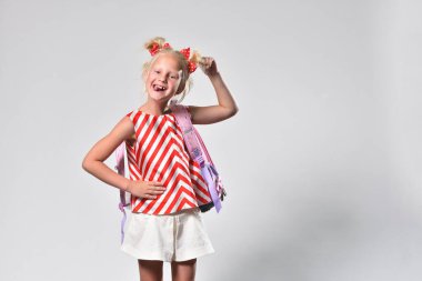 Sırt çantasıyla gülümseyen neşeli sarışın kız gri arka planda dilini gösteriyor..