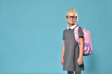 Ortaokula hazırlanan ve sırt çantasıyla mavi bir arka planda poz veren tatlı bir anaokulu kızı.