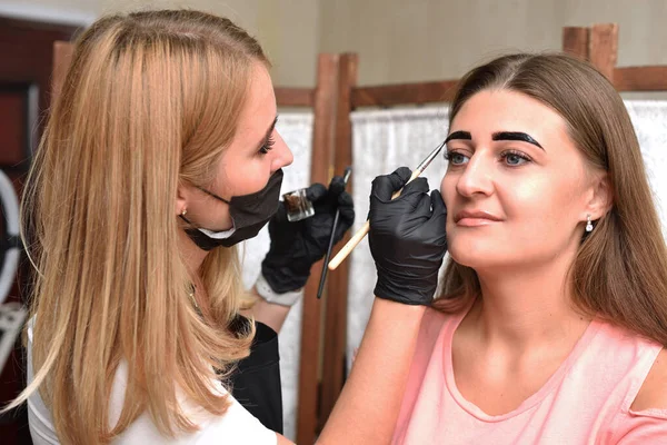 Beautician makes eyebrow coloring with henna to a young woman in a beauty salon