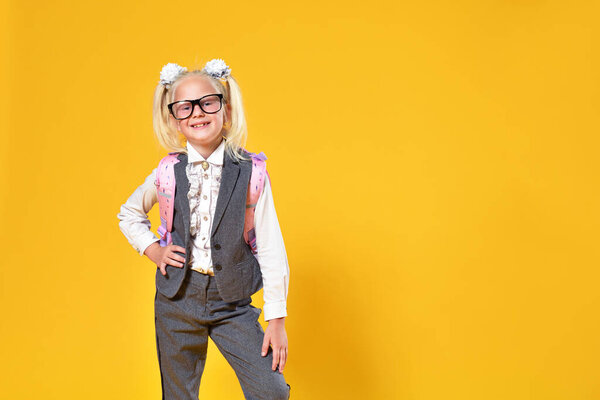 Young schoolgirl with glasses and a backpack smiling on a yellow background