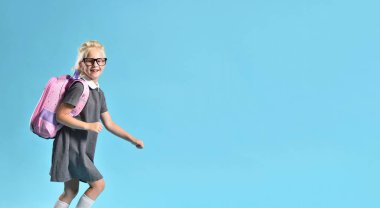 Sırt çantalı bir liseli kız mavi arka planda neşeli bir şekilde koşuyor.