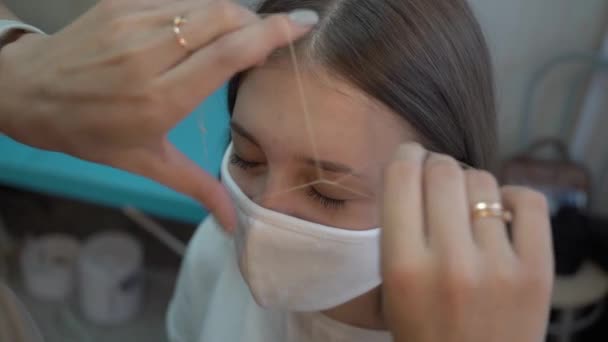 Chica joven en una máscara protectora arrancando las cejas con un hilo en un salón de belleza — Vídeos de Stock