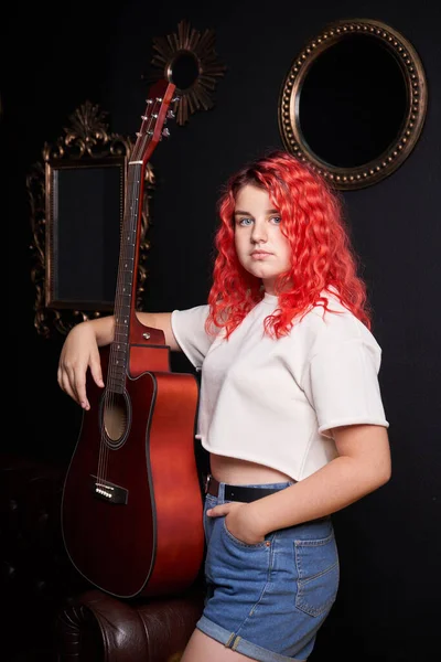 Retrato de uma adolescente com cabelo vermelho e uma guitarra acústica em um fundo preto — Fotografia de Stock