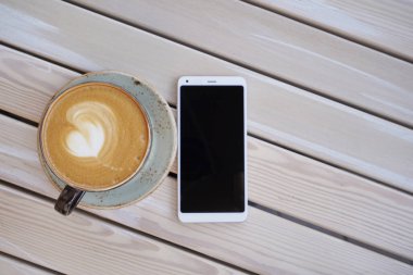 Aromatik kapuçino kahveli büyük bardak ve ahşap bir masada beyaz akıllı telefon.