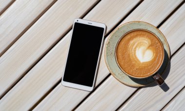 Bardakta nefis bir kahve ve bir kafede ahşap bir masada beyaz bir akıllı telefon.