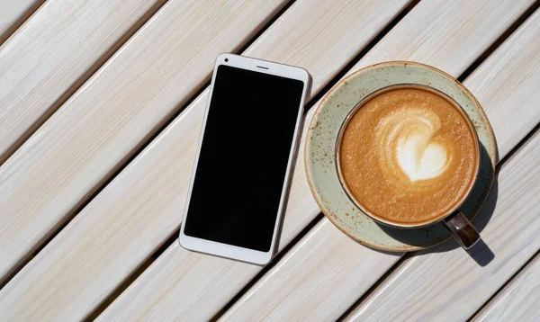 Bardakta nefis bir kahve ve bir kafede ahşap bir masada beyaz bir akıllı telefon.