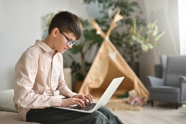 Adolescente menino com óculos funciona em seu laptop contra o fundo de seu quarto — Fotografia de Stock
