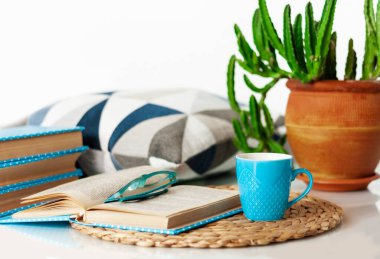 Konforlu ev dekorasyonu: bir fincan kahve, kitap yığını, saksıda bitkiler, hasır tezgahta açık kitap, beyaz bir masada yastıklar. Uzak ev eğitimi. Evde kalma karantina kavramı.