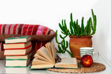 Konforlu ev dekorasyonu: bir fincan kahve, kitap yığını, saksıda bitkiler, açık kitap ve hasır tezgahta kırmızı şeftali, beyaz bir masada yastıklar. Uzak ev eğitimi. Evde kalma karantina kavramı.